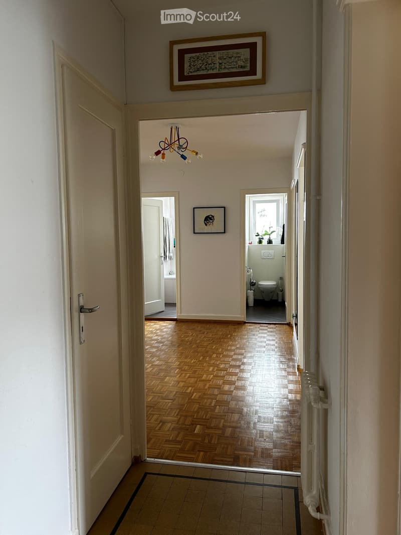 Hallway with wooden flooring, white walls, and a chandelier. Doors lead to a bathroom and another room. Artwork is displayed on the walls.