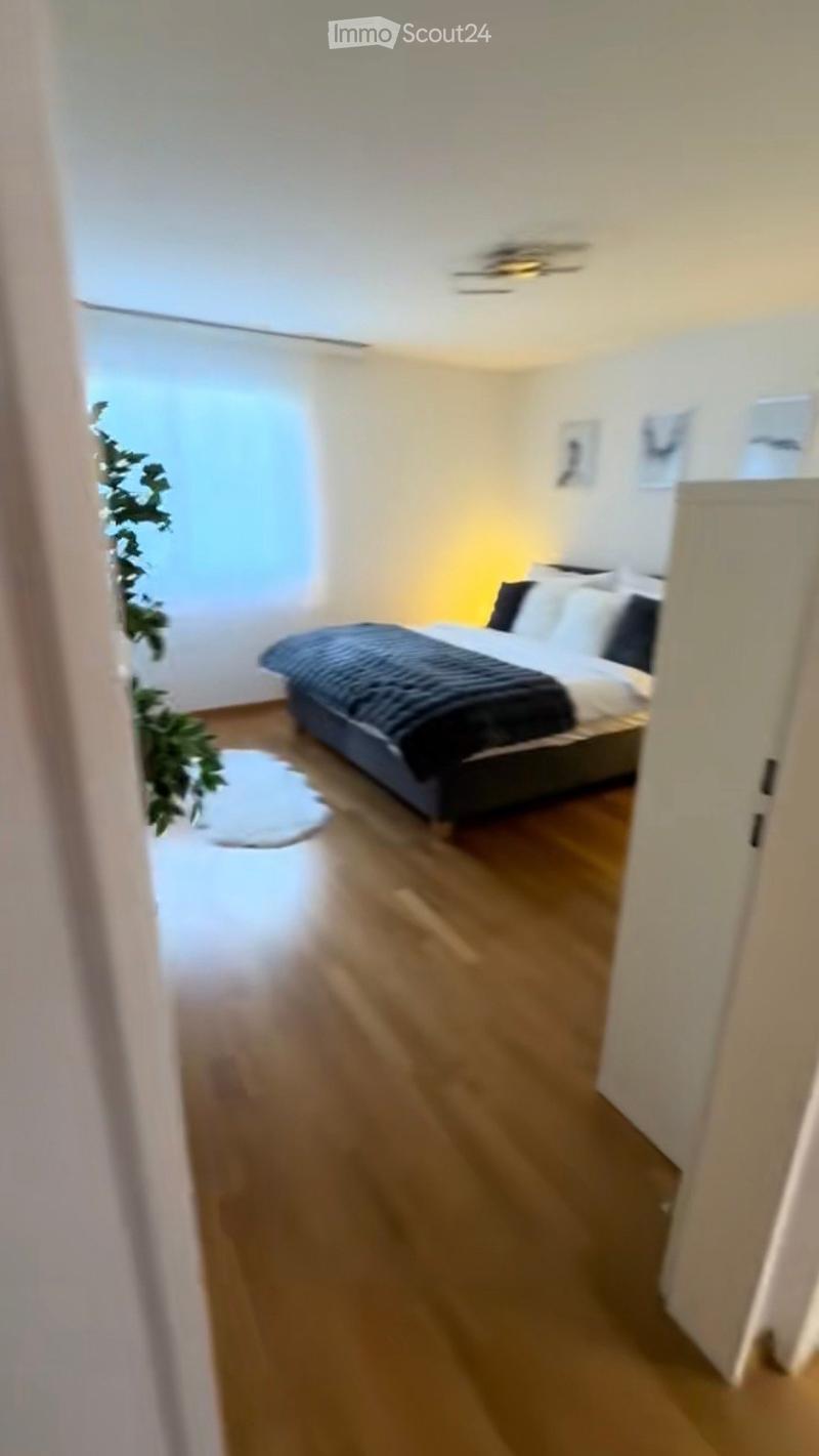 Modern bedroom with wooden flooring, a bed with dark bedding, a white rug, and a large window with sheer curtains. The room features a ceiling light fixture and a small plant near the window.