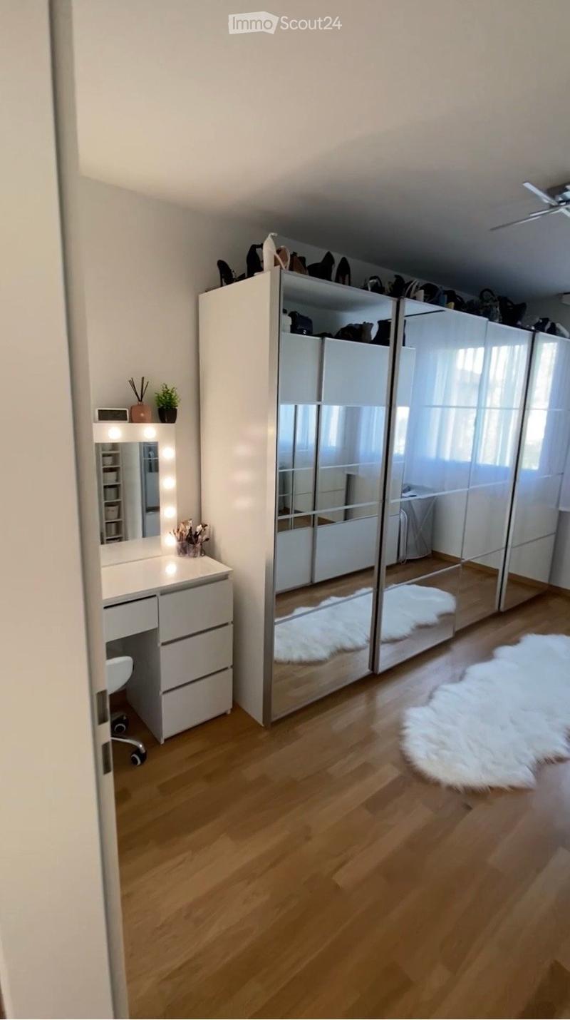 A well-lit, modern walk-in closet with a large mirrored wardrobe, a vanity desk with a mirror, a small plant, and a white fur rug on the wooden floor.