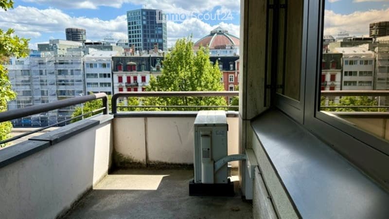 Balcony with metal railing, overlooking a cityscape with buildings and trees. A small air conditioning unit is placed on the floor.