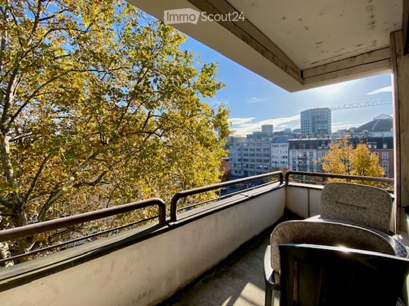 Balcony with metal railing, overlooking cityscape and autumn trees, with a single chair placed on it.