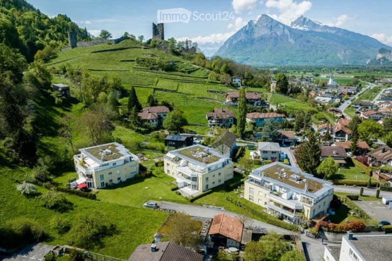 Aerial view of a village nestled in a lush green valley with mountains in the background. The village features a mix of residential buildings, including modern apartments and traditional houses, surrounded by fields and trees.