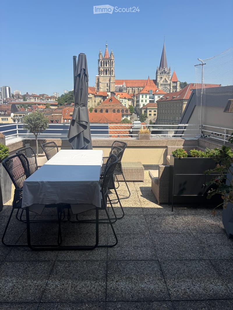 Rooftop terrace with a table, chairs, sofa, and umbrella, overlooking a cityscape with historic buildings.