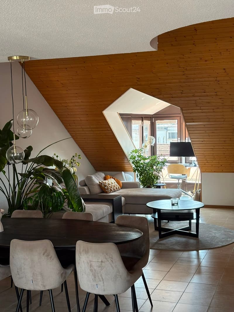 Modern living room with a slanted wooden ceiling, large window, sofa, coffee table, dining table, chairs, and decorative plants.