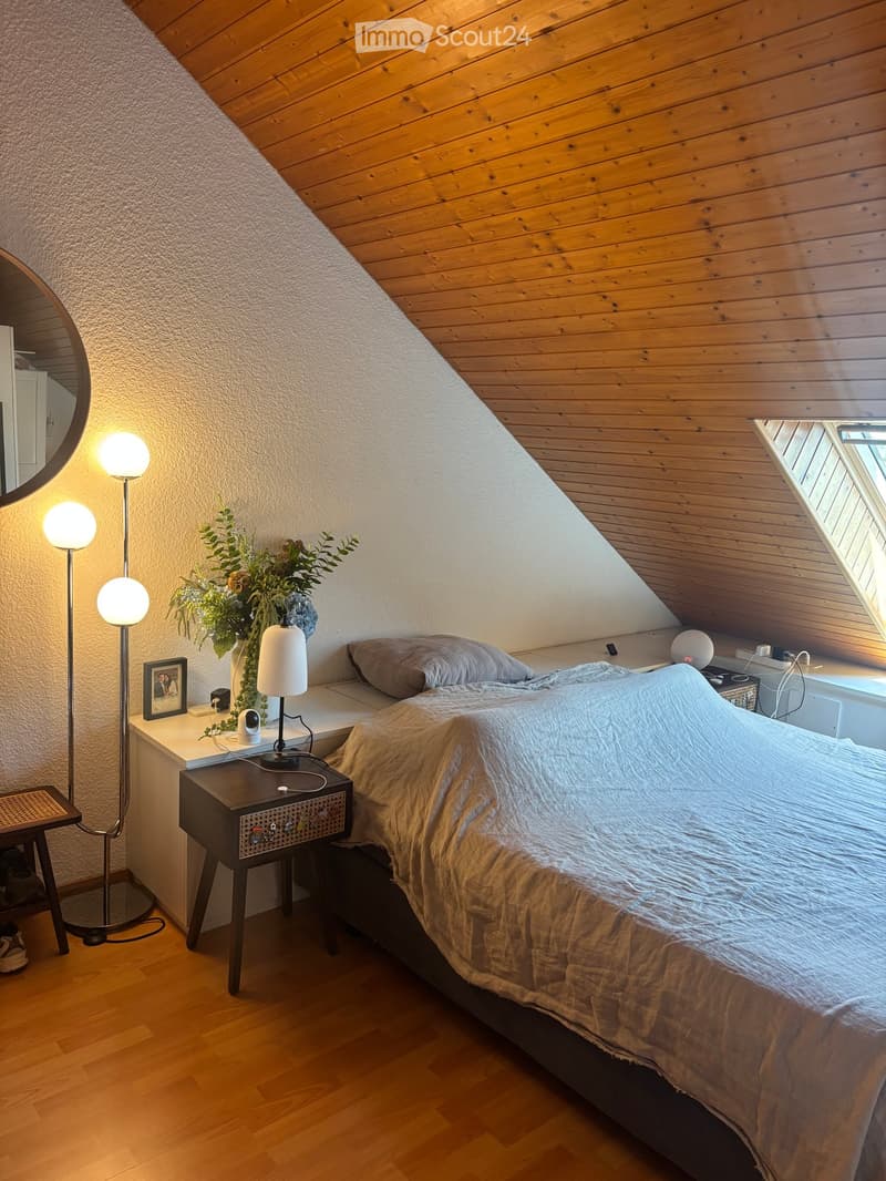 Cozy attic bedroom with wooden slanted ceilings, a large bed with a light-colored duvet, a nightstand with a lamp, a mirror, and a vase with flowers on a small table.