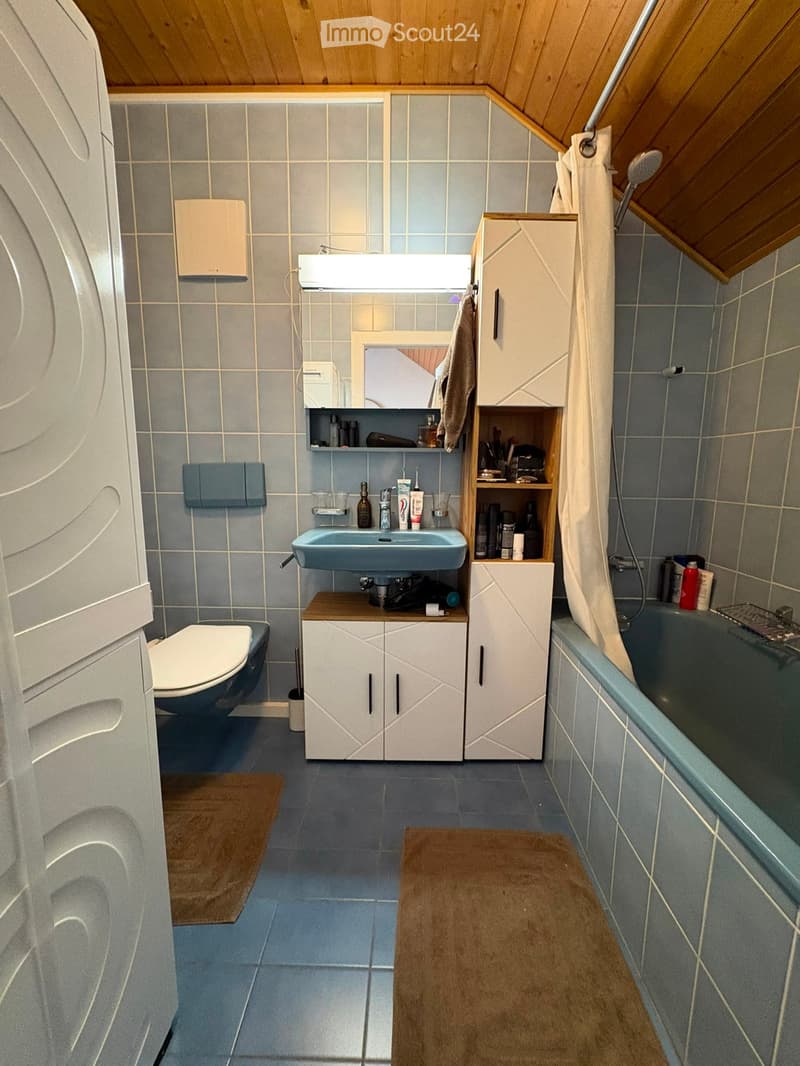 A small, well-organized bathroom with a toilet, sink, bathtub, and built-in cabinets. The room features light blue tiles on the walls and floor, a wooden ceiling, and a small wooden shelf holding various toiletries.