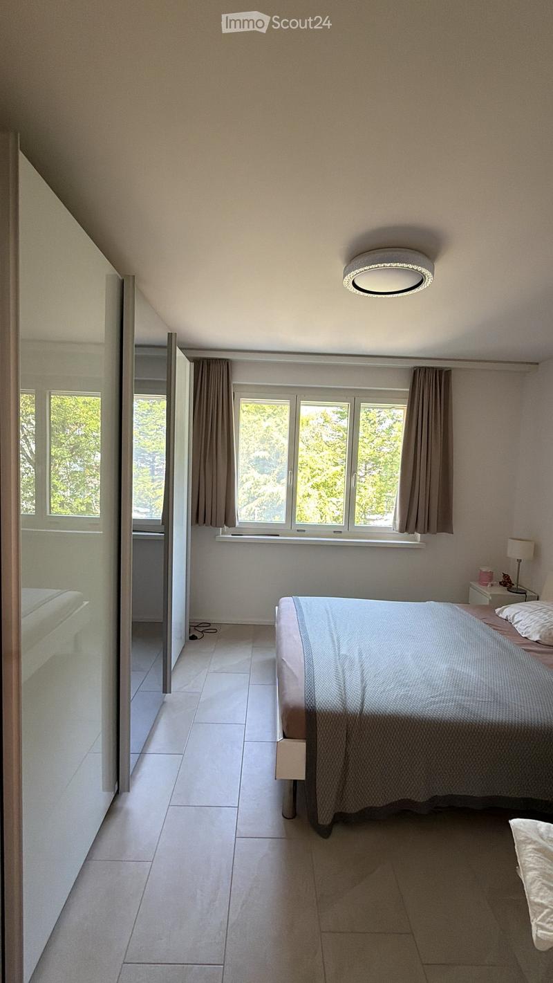 Modern bedroom with a large bed, nightstand, lamp, and large windows with curtains. The room features a sleek, minimalist design with a skylight ceiling fixture.