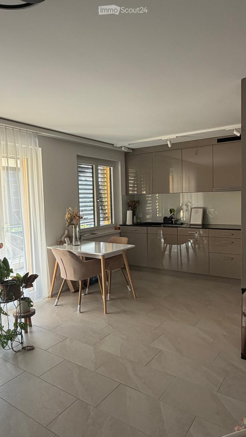 Modern kitchen with light grey cabinets, a white dining table with four chairs, a sink, and decorative items including a framed picture and vases. The space features large windows with blinds and a tiled floor.