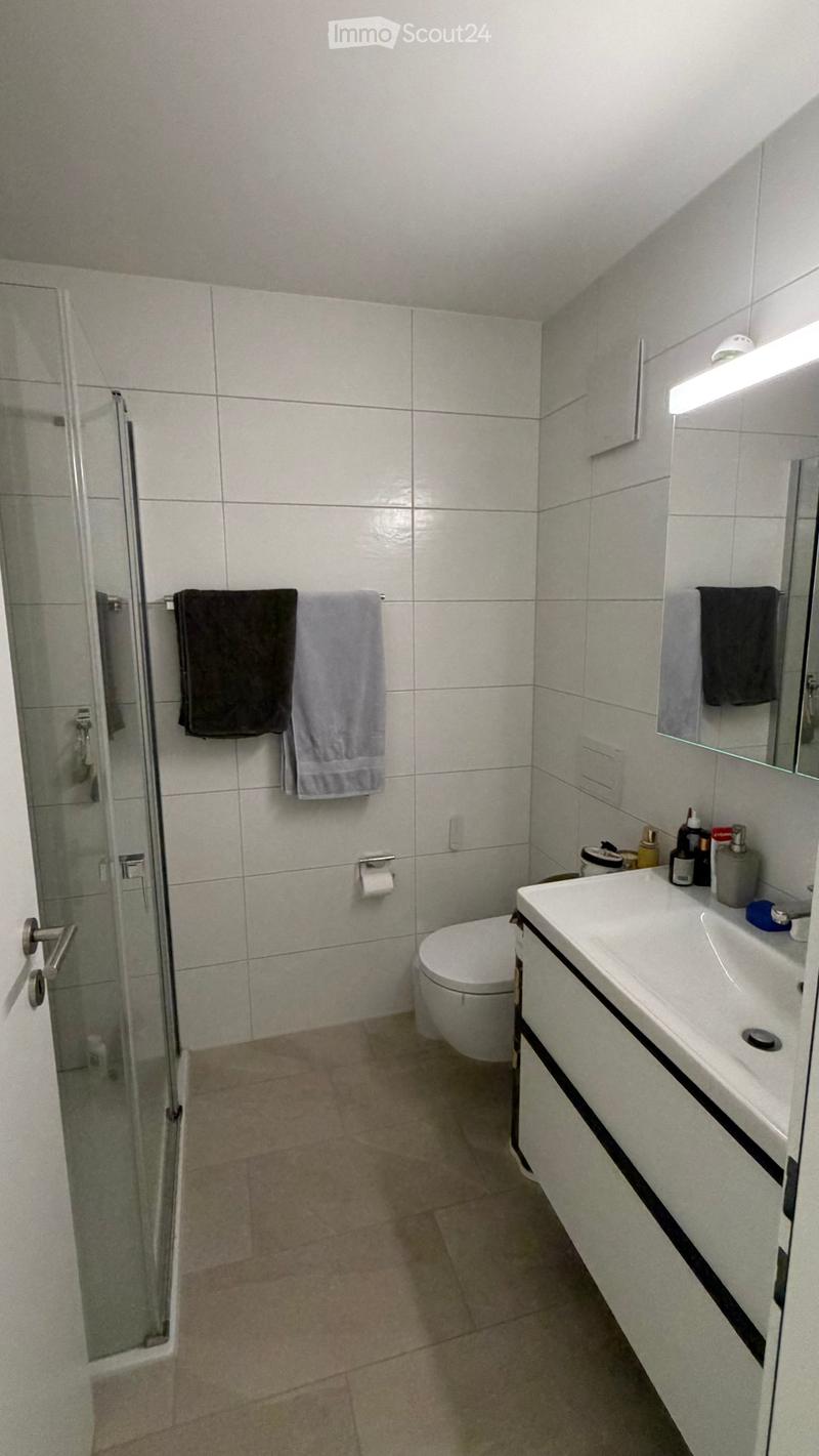 Modern bathroom with white tiles, a toilet, a sink with a mirror, a glass-enclosed shower, and towels hanging on a rack.