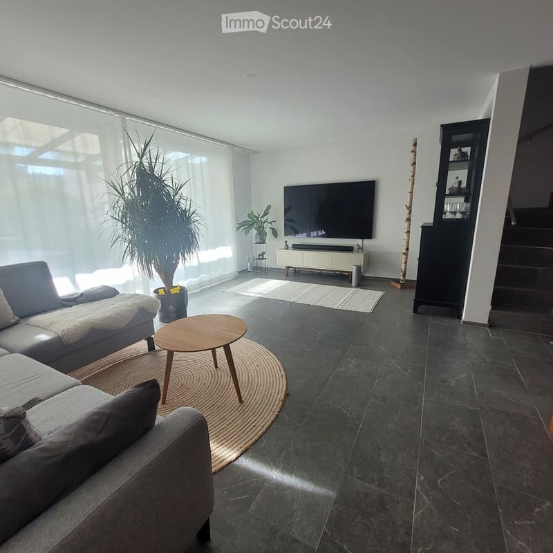 Modern living room with grey sectional sofa, wooden coffee table, large flat-screen TV, and indoor plants. The space features dark tiled flooring and large windows with sheer curtains.
