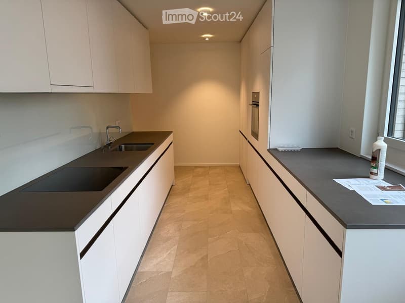 Modern kitchen with white cabinets, dark countertops, and a built-in sink and oven. A bottle is placed on the counter near the window.