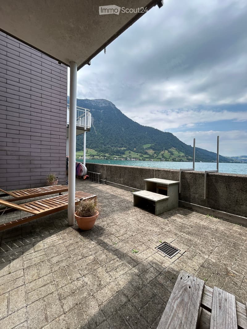 Balcony with stone tiles, wooden sun loungers, a small table and chairs, and potted plants, overlooking a scenic lake and mountain view.