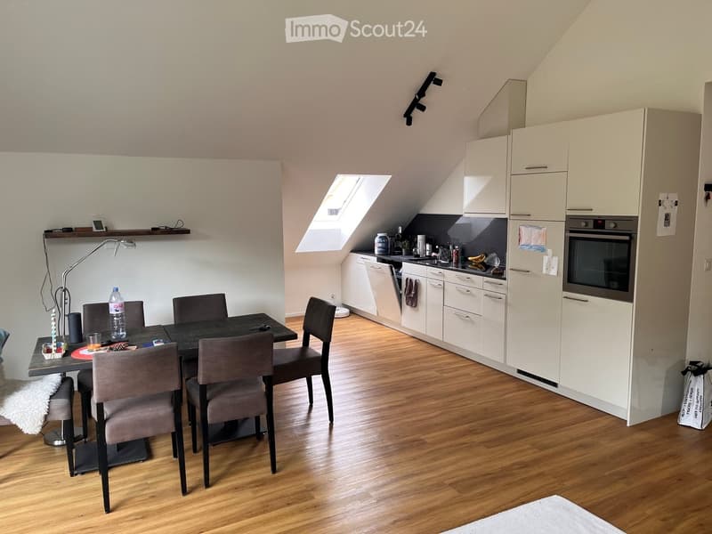 A modern, compact kitchen and dining area with white cabinets, a black dining table, four chairs, and various kitchen appliances including an oven, dishwasher, and refrigerator. The space features wooden flooring and a skylight window providing natural li