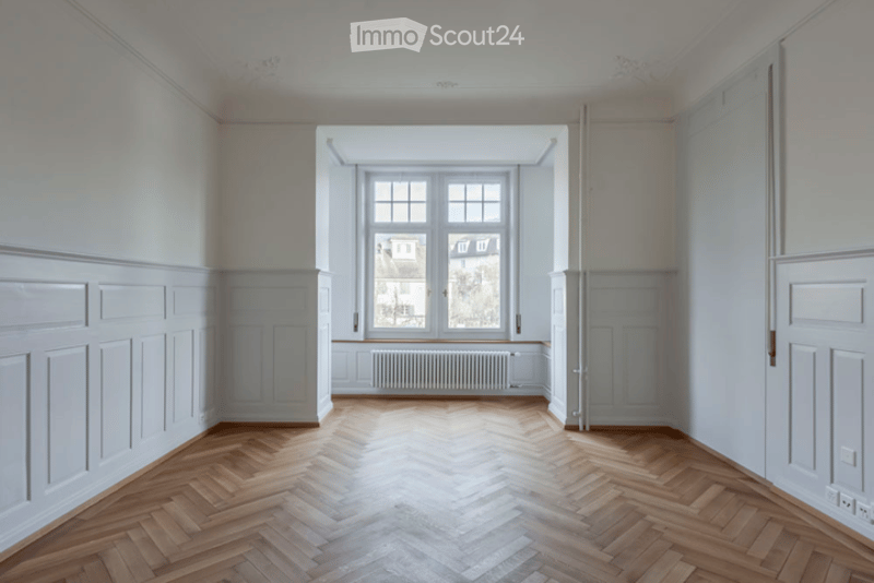Empty room with wooden herringbone flooring, white walls, large window with radiator below, and white paneling on walls.
