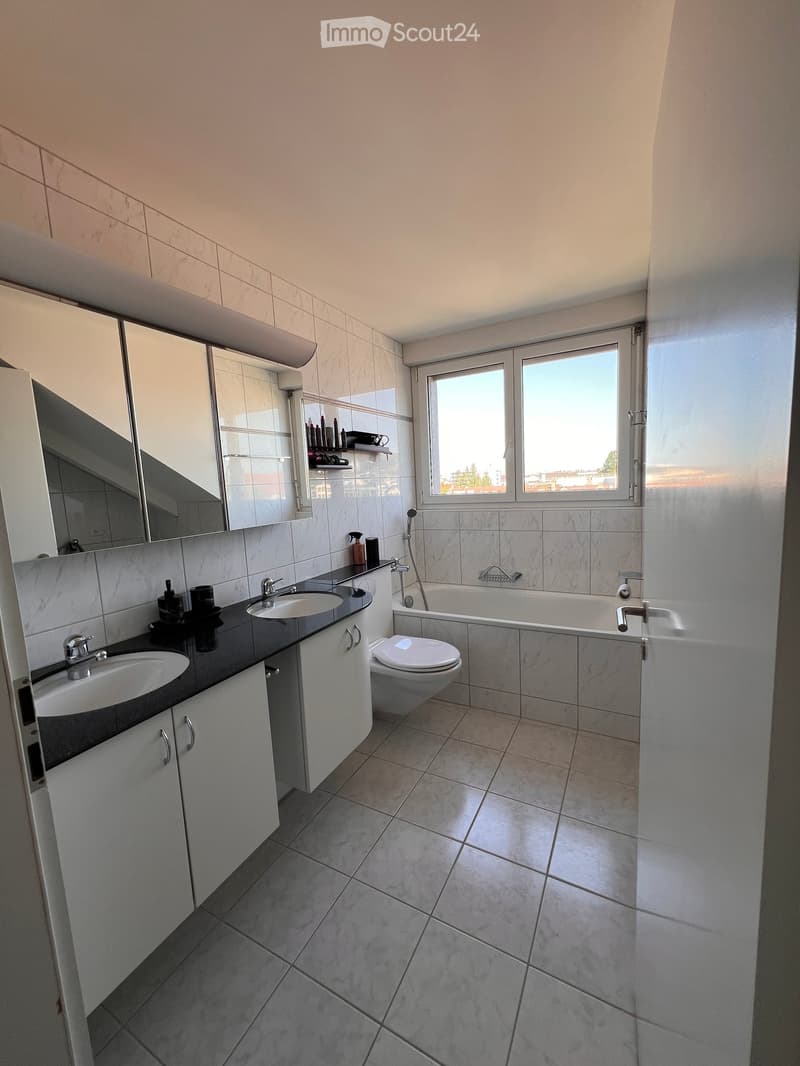 Modern bathroom with two sinks, a toilet, a bathtub, and a large mirror above the sinks. The room features light-colored tiles and a window providing natural light.