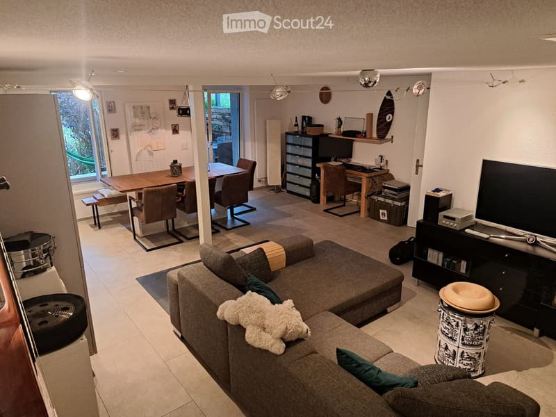 Open-plan living and dining area with a grey sectional sofa, wooden dining table, chairs, a laptop on a desk, and a TV on a stand.
