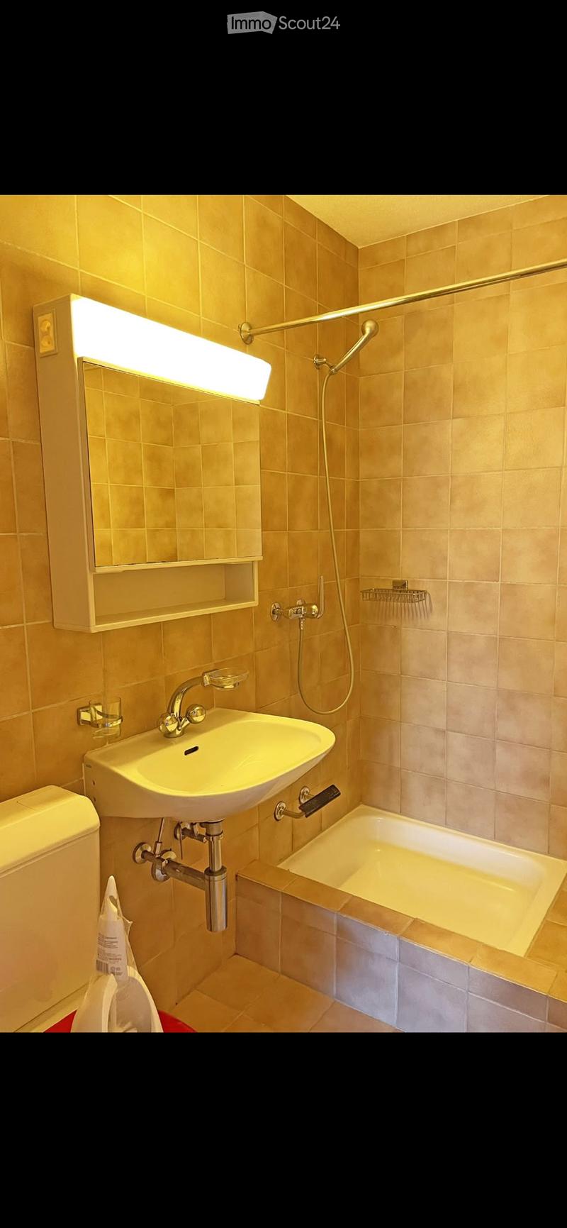 Bathroom with beige tiles, a white sink with a chrome faucet, a bathtub with a shower hose, a mirror with built-in lighting, and a toilet.