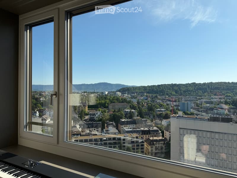 View from a window showing a cityscape with buildings, trees, and distant hills under a clear blue sky.