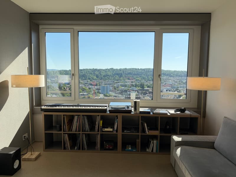 Modern living room with large windows offering a scenic city view, featuring a grey sofa, a wooden bookshelf filled with books and records, a record player, a keyboard, and two floor lamps.