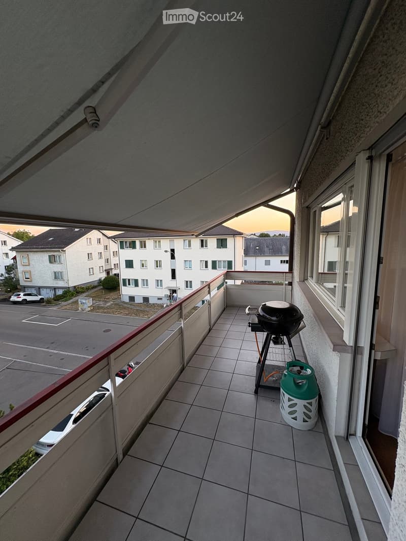 Balcony with tiled floor, metal railing, grill, and gas tank. View of parking lot and residential buildings.