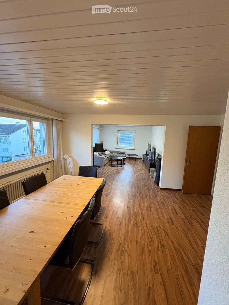 Bright, open-plan living and dining area with wooden flooring, large windows, and modern furniture including a dining table, chairs, sofa, TV, and lamp.