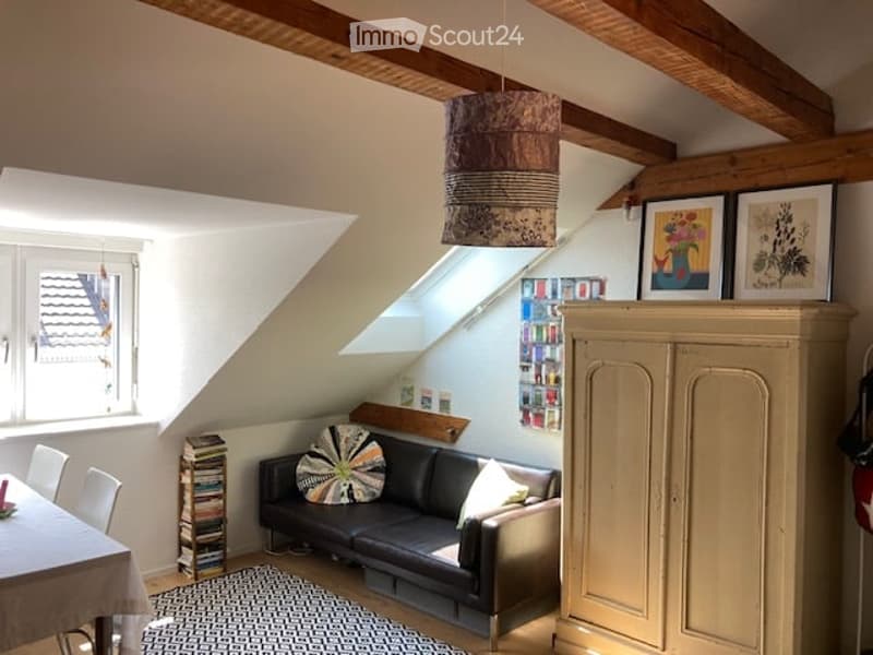 Cozy attic room with slanted wooden ceiling beams, exposed wooden beams, large skylight window, black leather sofa, colorful throw pillow, small dining table with chairs, books stacked on floor, and a tall cabinet with picture frames on top.
