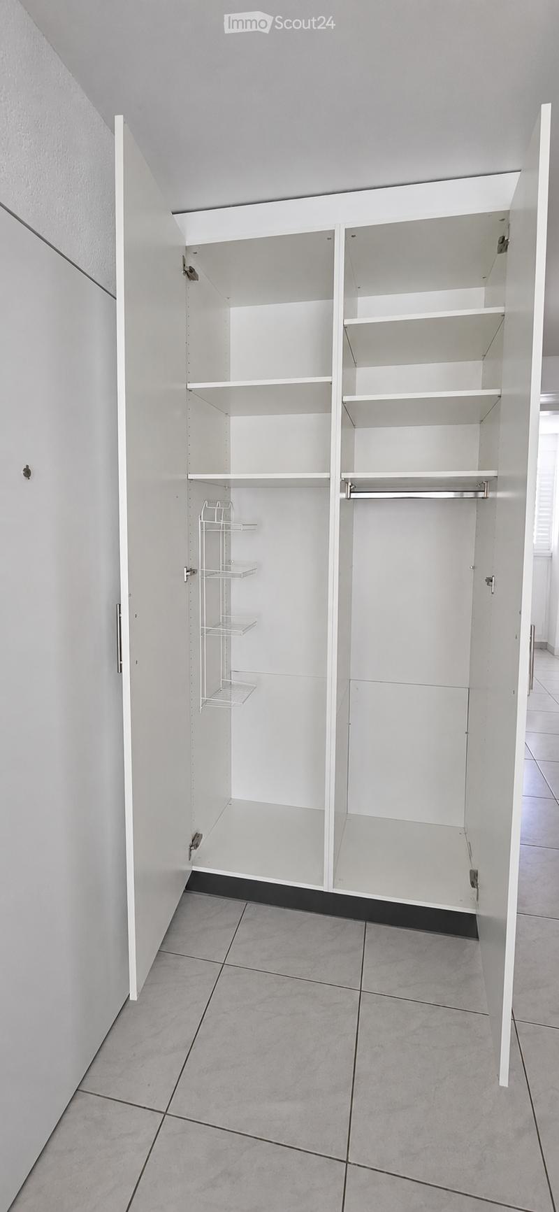 Empty closet with white built-in cabinets and shelves, featuring a hanging rod and wire basket, set in a tiled room with light-colored walls.