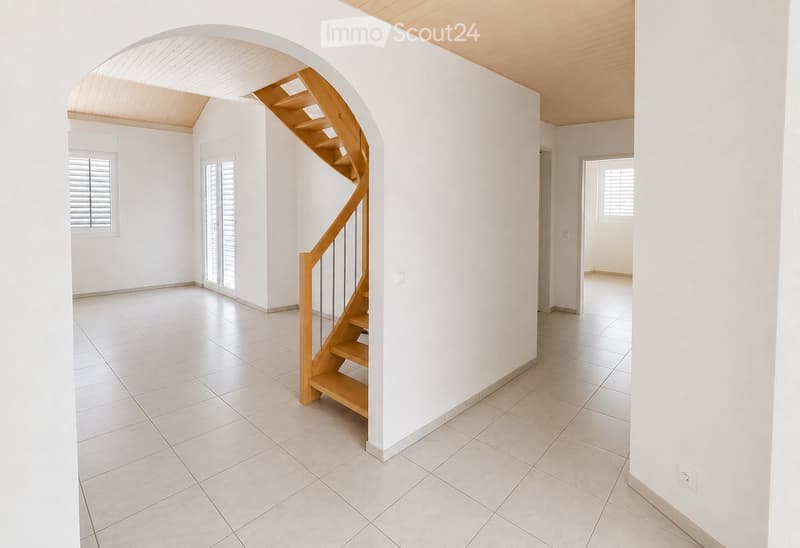 Empty hallway with white tiled flooring, white walls, and a wooden staircase leading upwards. Features include a large arched opening, a window with shutters, and a door leading to another room.