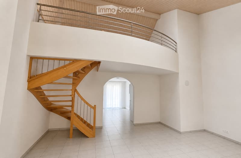Spacious hallway with a wooden staircase leading to an upper level, white walls, tiled floor, and a balcony railing visible above.