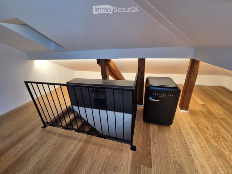 An attic space with wooden flooring, exposed wooden beams, and a staircase with a safety railing. A small black cabinet and a black mini-fridge are present.
