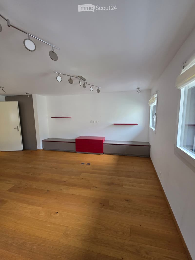 Empty room with wooden flooring, white walls, and modern lighting fixtures. Features a built-in cabinet and a small red shelf.