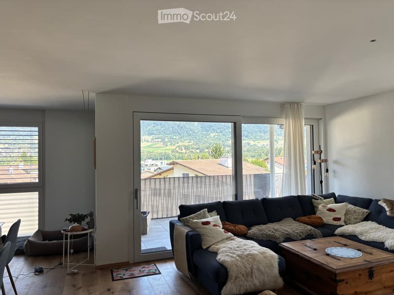 Modern living room with large windows offering a scenic view, a dark blue sectional sofa with multiple throw pillows and a furry throw, a wooden coffee table, a side table with a plant, and a small dog bed.
