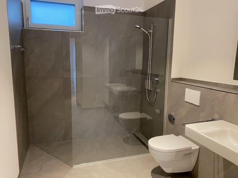 Modern bathroom with a glass-enclosed shower, toilet, and sink. The walls are tiled in a neutral gray color, and the floor is tiled in a light beige color. There is a small window above the shower for natural light.