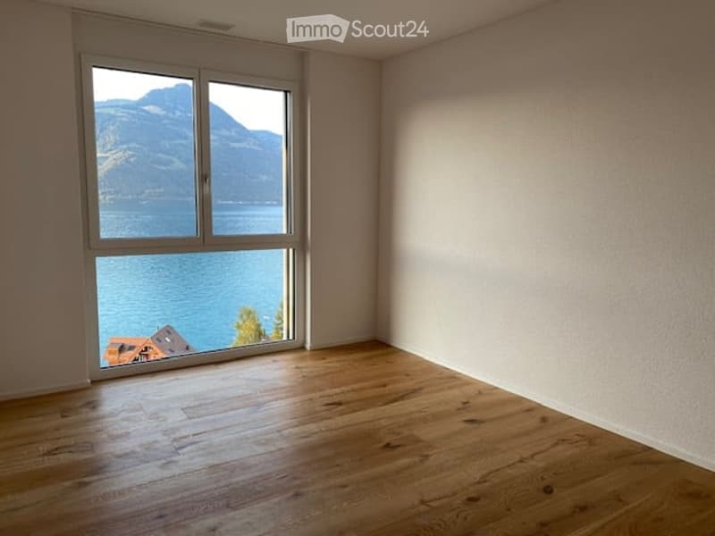 Empty room with wooden flooring and large window offering a scenic view of a lake and mountains.