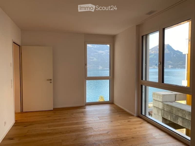 Empty room with wooden flooring, large windows offering a scenic view of a lake and mountains, and a closed door.