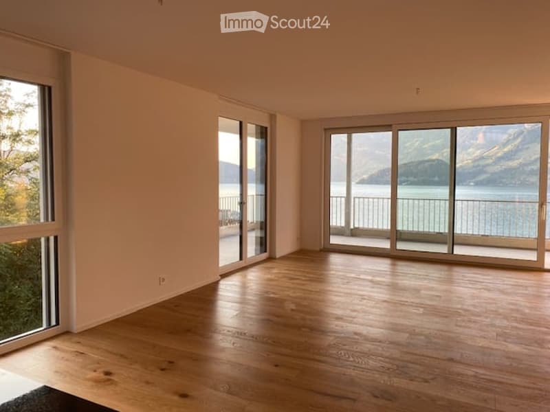 Bright, empty room with large windows and sliding glass doors leading to a balcony overlooking a scenic lake and mountains. The floor is made of polished wood.