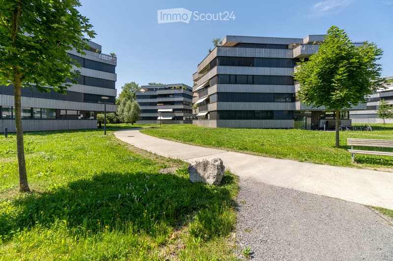 Modern residential complex with contemporary buildings, green lawn, pathways, trees, and a bench.