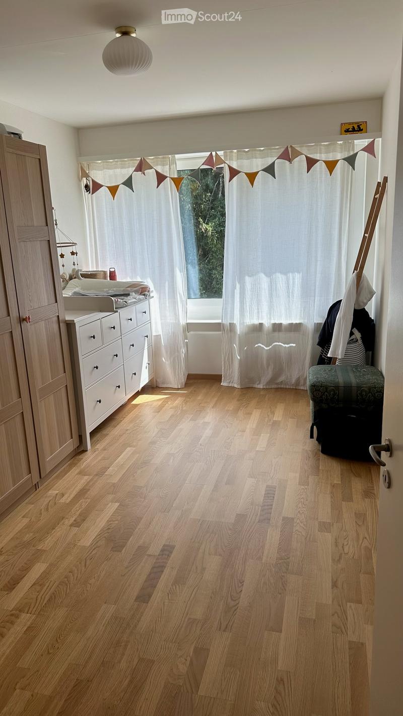 A well-lit room with wooden flooring, a large window with white curtains, a wardrobe, a drawer unit, a small stool, and a decorative banner above the window.