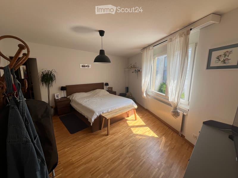 Bright bedroom with wooden flooring, large bed with white bedding, nightstand, bench at the foot of the bed, and large windows with white curtains.