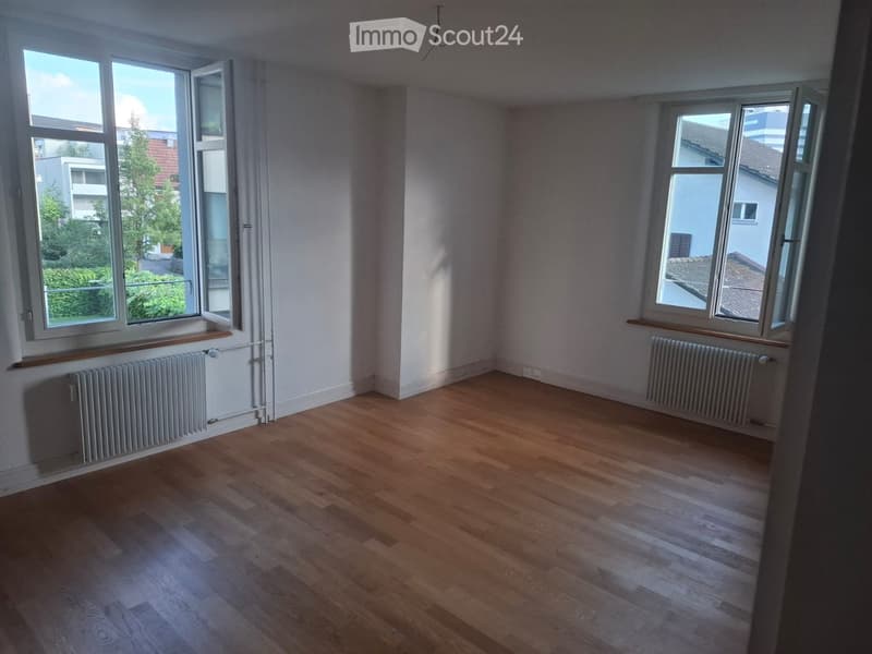Empty room with wooden flooring, white walls, and large windows with radiators beneath them.