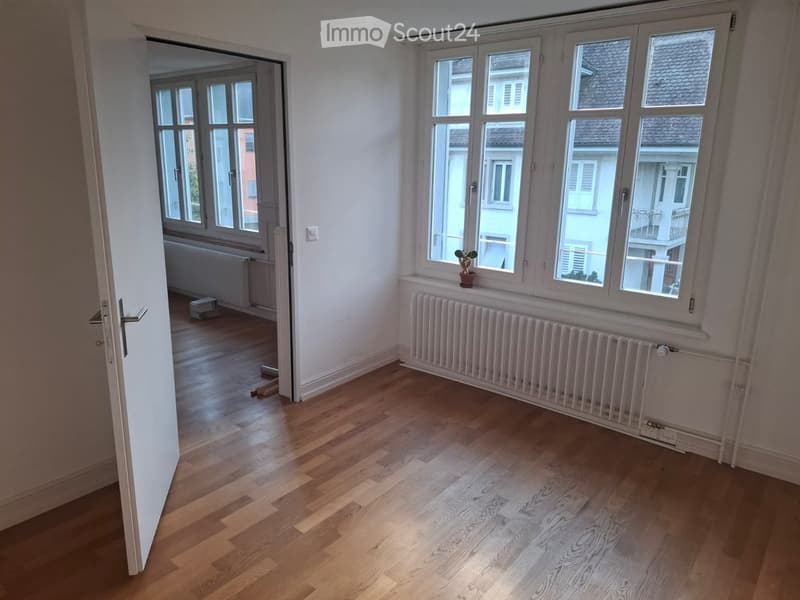 Empty room with wooden flooring, large windows, radiators, and a small potted plant on the windowsill.