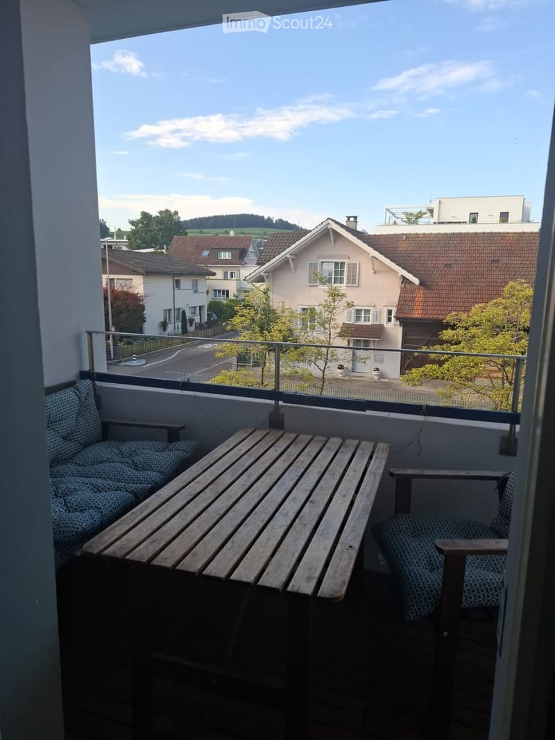 Balcony with wooden table and two chairs, overlooking a residential area with houses and trees.