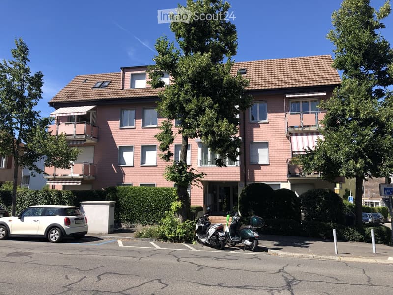 Two-story house with pink brick facade, multiple windows, balconies, and a tiled roof. The house is surrounded by greenery and has a small entrance area with steps.