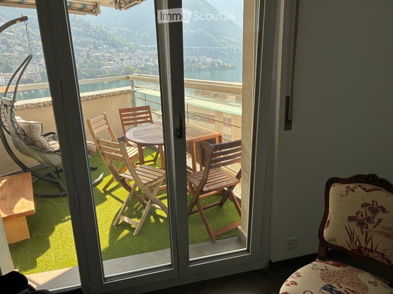 Balcony with a scenic view of a lake and mountains, featuring a round wooden table, four wooden chairs, and a hanging wicker chair.