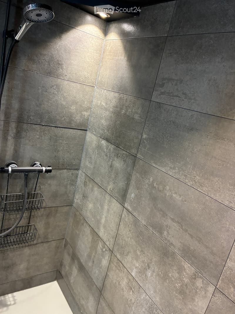 Modern shower area with grey tiled walls, a handheld showerhead, a shower rail, and a small metal basket for storage.