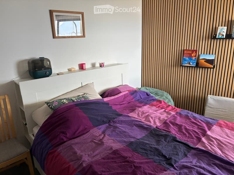 A cozy bedroom with a bed covered in a colorful patchwork quilt, a wooden chair, a mirror on the wall, a humidifier on the nightstand, and a few decorative items including books and a cup.