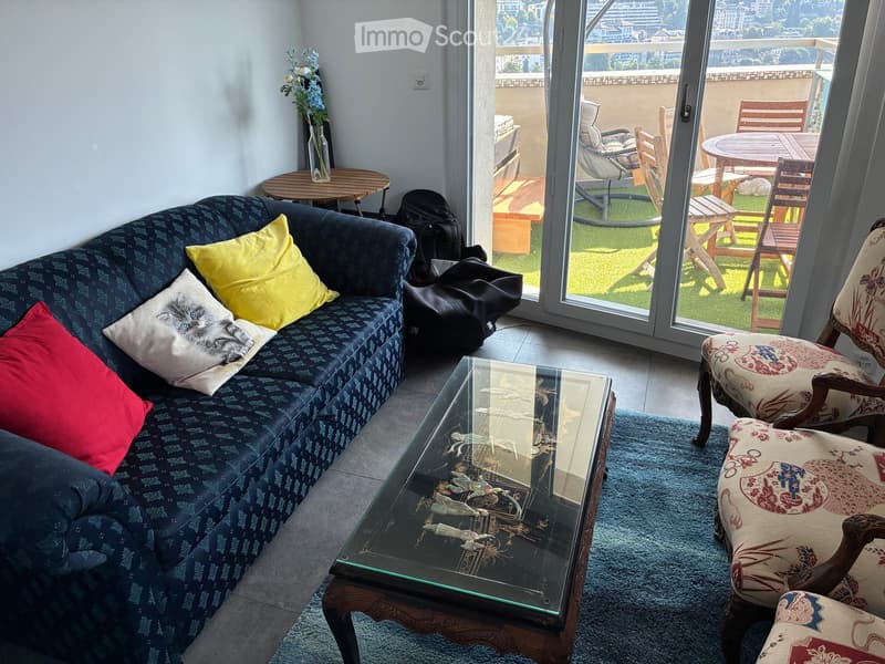 Living room with a blue sofa, glass coffee table, patterned chairs, and a small table with a vase of flowers. A balcony with outdoor furniture is visible through large glass doors.