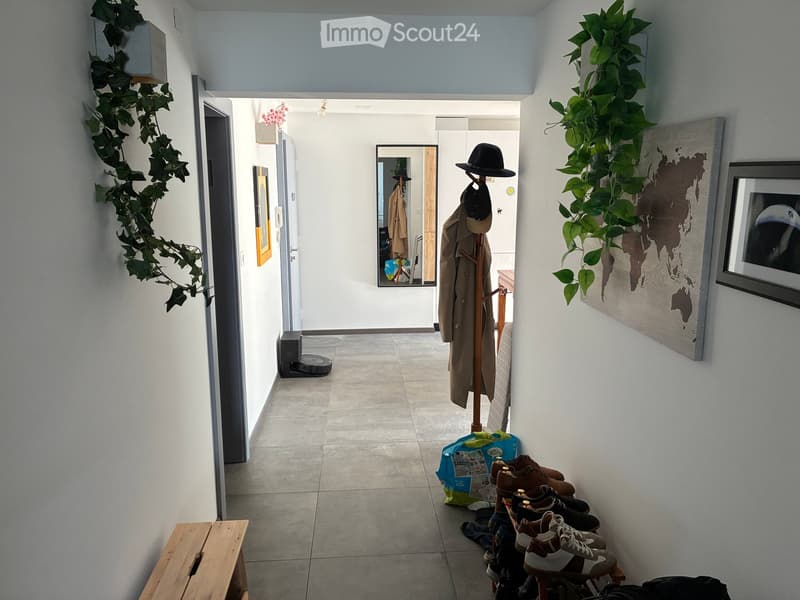 A well-lit hallway with white walls, tiled floor, and decorative elements including a coat rack with coats and hats, a shoe rack with multiple pairs of shoes, a small wooden bench, and wall-mounted plants. A mirror reflects part of the adjacent room.