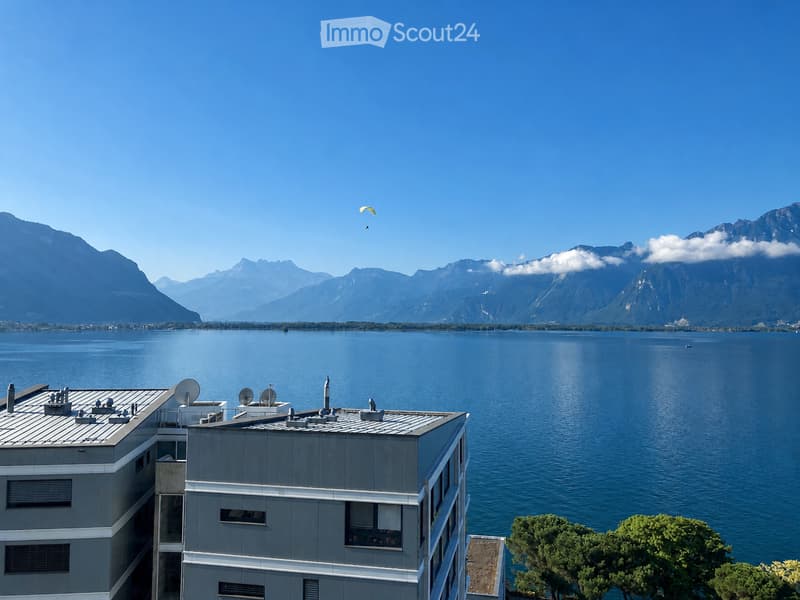 Modern multi-story building with grey facade and white trim, overlooking a large lake and mountain range under a clear blue sky.