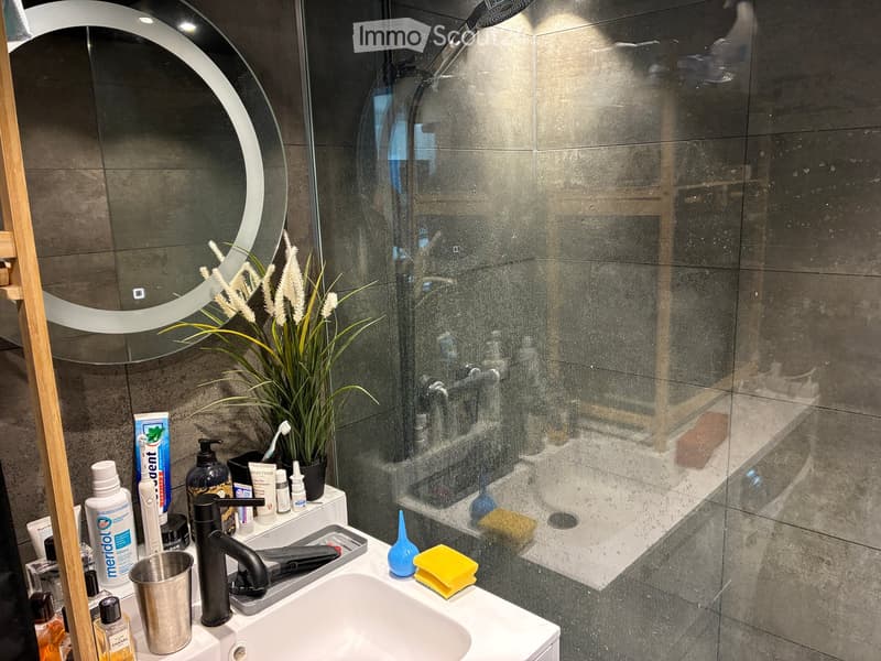 Modern bathroom with a white sink, black faucet, round mirror, and a glass-enclosed shower. The countertop holds various toiletries, a plant, and a yellow sponge.